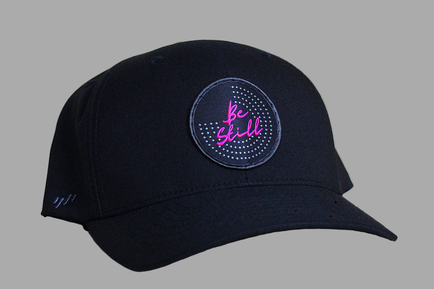 Black cap with a circular patch featuring 'Be Still' in pink text on a gray background