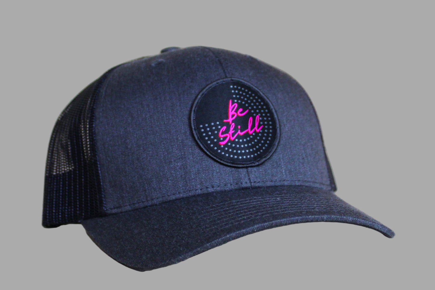 Navy blue cap with pink 'Be Still' patch on a gray background
