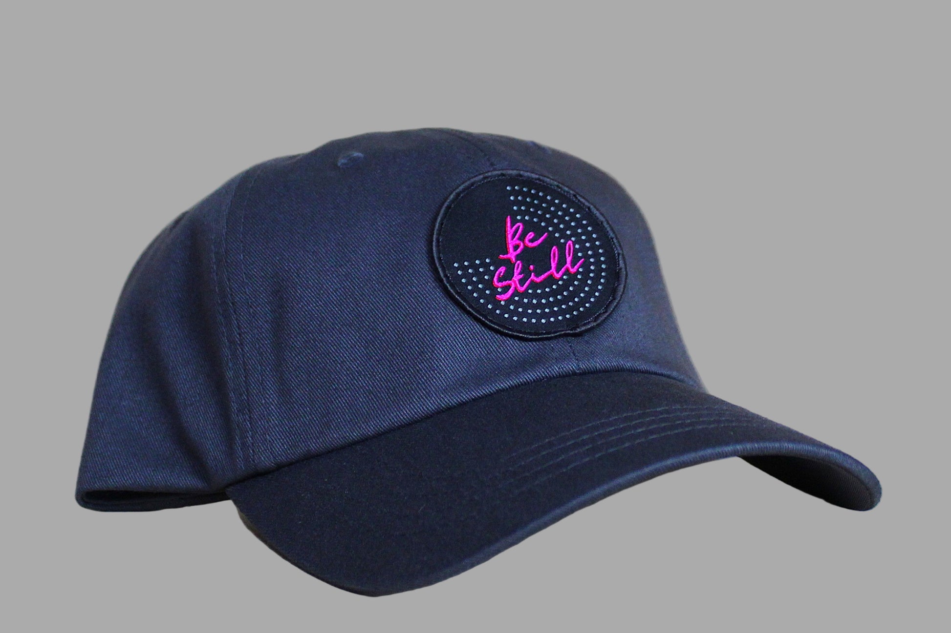 Navy blue cap with a circular patch featuring 'Be Still' in pink text on a light wooden surface.