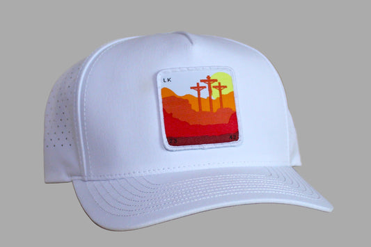 White cap with a colorful patch featuring crosses and a sunset on a wooden surface